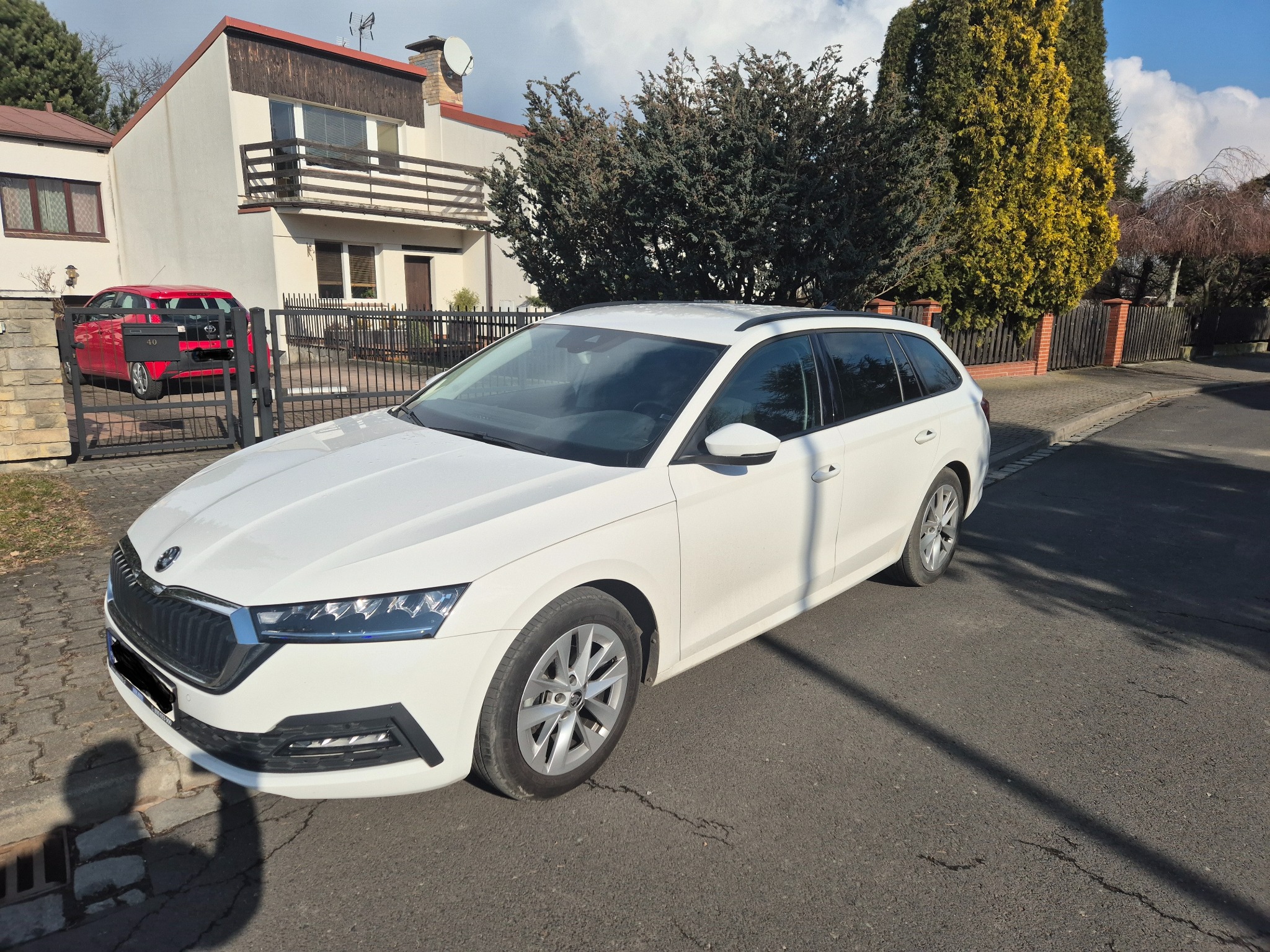 Biała Skoda Octavia Combi zaparkowana na ulicy przed domem jednorodzinnym z czerwonym autem w tle.