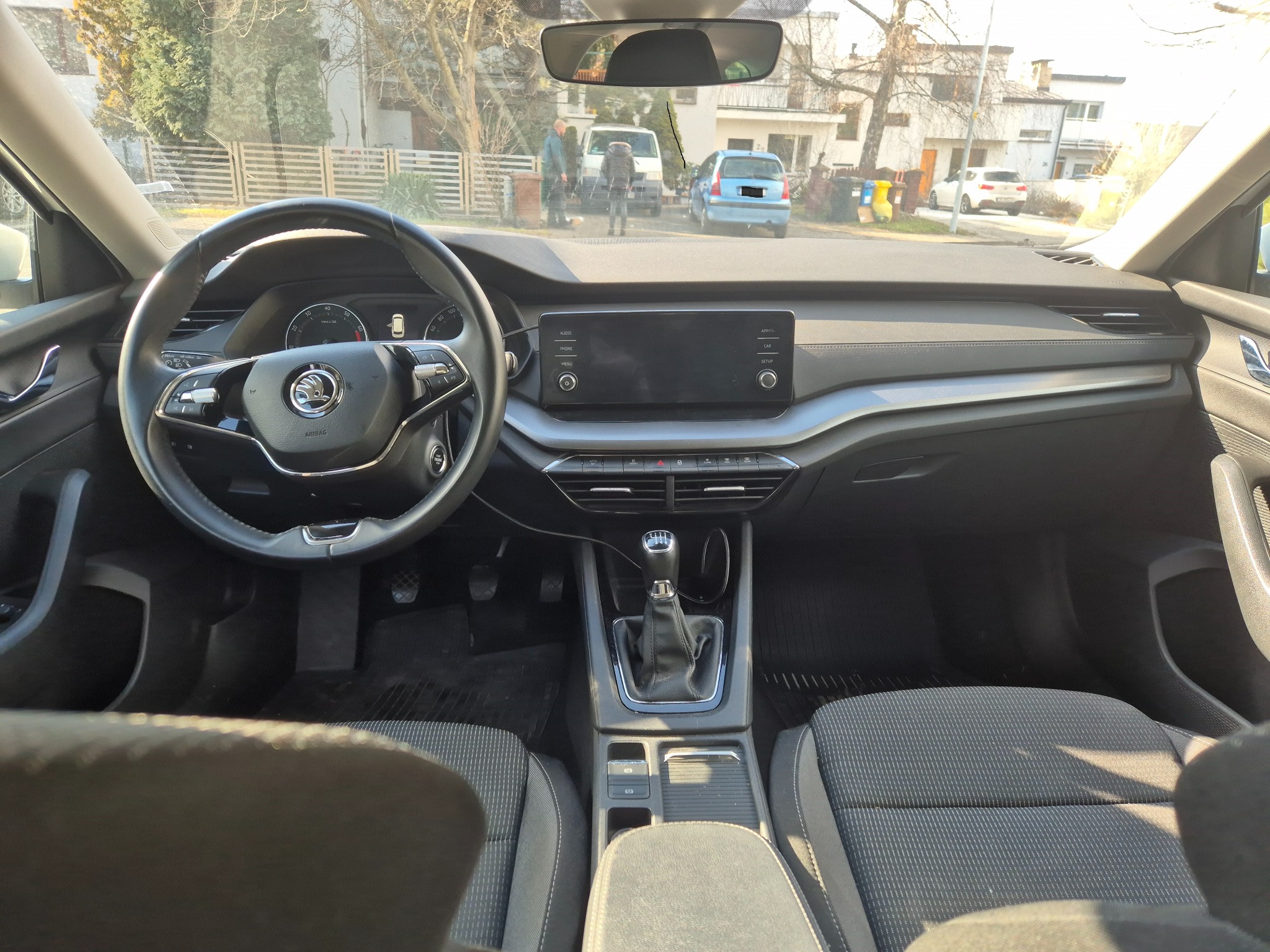 Wnętrze samochodu osobowego marki Skoda z manualną skrzynią biegów, widok z perspektywy pasażera na deskę rozdzielczą, kierownicę i ulicę z budynkami w tle.