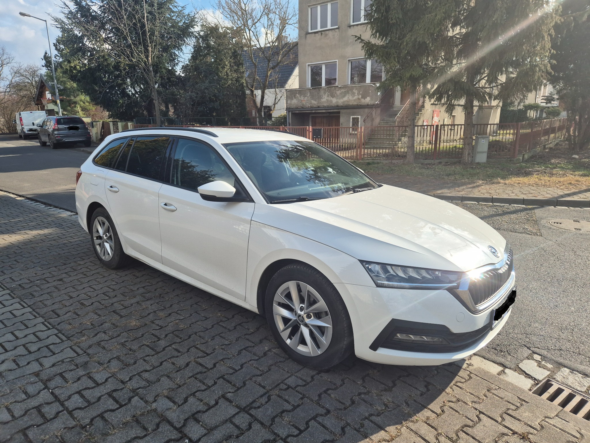 Biała Skoda Octavia kombi zaparkowana na brukowanej ulicy w słoneczny dzień, widoczny budynek mieszkalny w tle.