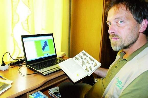 Mężczyzna z brodą siedzi przy biurku, obok laptop z otwartą stroną z ilustracją ptaka, w rękach trzyma otwartą książkę z rysunkami ptaków.