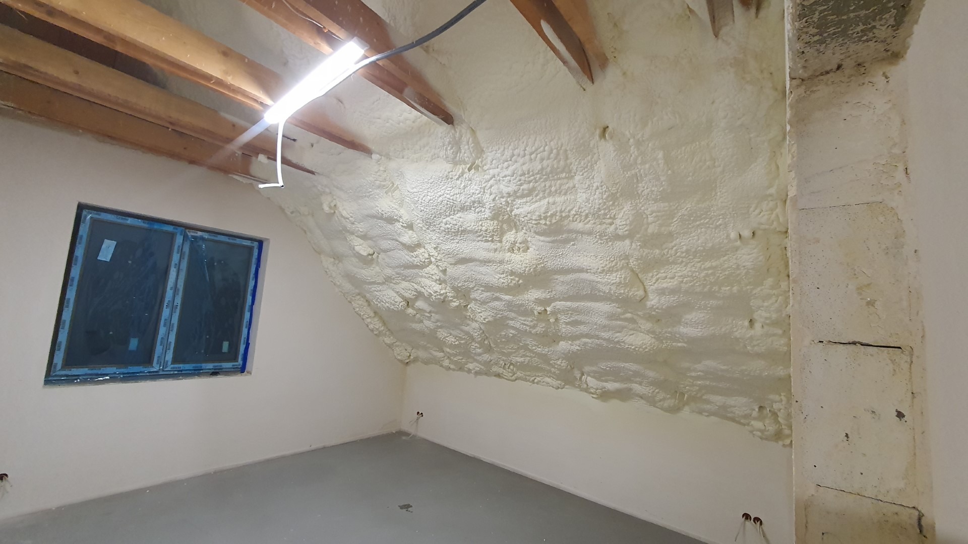 Poddasze po termoizolacji, ściany i skosy pokryte jasną pianą izolacyjną, widoczne drewniane belki stropowe i okno z niebieską folią ochronną, świeżo wykończone wnętrze w Gliwicach.