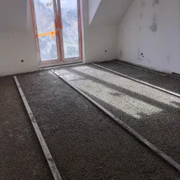 Świeżo wylana posadzka z widocznymi łatami poziomującymi w jasnym pomieszczeniu na poddaszu z oknem zabezpieczonym folią.