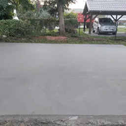 Świeżo wylana, gładka, szara posadzka na zewnątrz, z widocznym samochodem i zadaszeniem w tle.