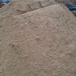 Kopa jasnego piasku z widocznym zagłębieniem na szczycie, w tle budynek w trakcie budowy z zielonym dachem.