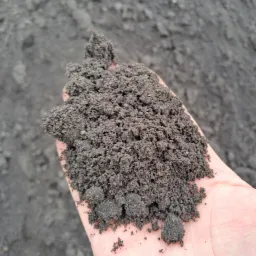 Garść ciemnej, wilgotnej ziemi w dłoni, na tle większej pryzmy tego samego materiału.