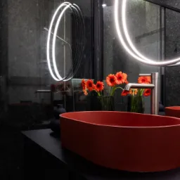 Nowoczesna łazienka z umywalką w kolorze terakoty, okrągłym lustrem z podświetleniem LED i szklaną kabiną prysznicową z deszczownicą.