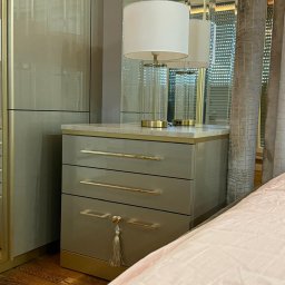 Wojciech Blarowski Meble Blarowscy - Szara szafka nocna z trzema szufladami i złotymi uchwytami, zwieńczona marmurowym blatem, na którym stoi szklana lampa z białym abażurem. Na jednej z szuflad wisi ozdobny chwost.