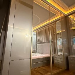 Wojciech Blarowski Meble Blarowscy - Nowoczesna szafa z przesuwnymi lustrzanymi drzwiami w sypialni, z widocznym łóżkiem z tapicerowanym zagłówkiem i oświetleniem LED na suficie.