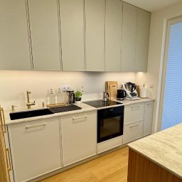 Wojciech Blarowski Meble Blarowscy - Nowoczesna kuchnia w odcieniach beżu i szarości z drewnianymi akcentami, czarnym zlewem i blatem z marmurkowym wzorem, oświetlona ledowym panelem.