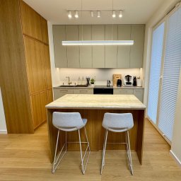 Wojciech Blarowski Meble Blarowscy - Nowoczesna kuchnia z drewnianymi szafkami, szarymi górnymi szafkami, wyspą z blatem z jasnego kamienia i dwoma szarymi krzesłami barowymi na białych nogach.