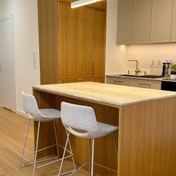 Wojciech Blarowski Meble Blarowscy - Nowoczesna kuchnia z drewnianą zabudową, wyspą z blatem w odcieniach beżu i dwoma szarymi krzesłami barowymi na metalowych nogach, oświetlona podłużną lampą LED.