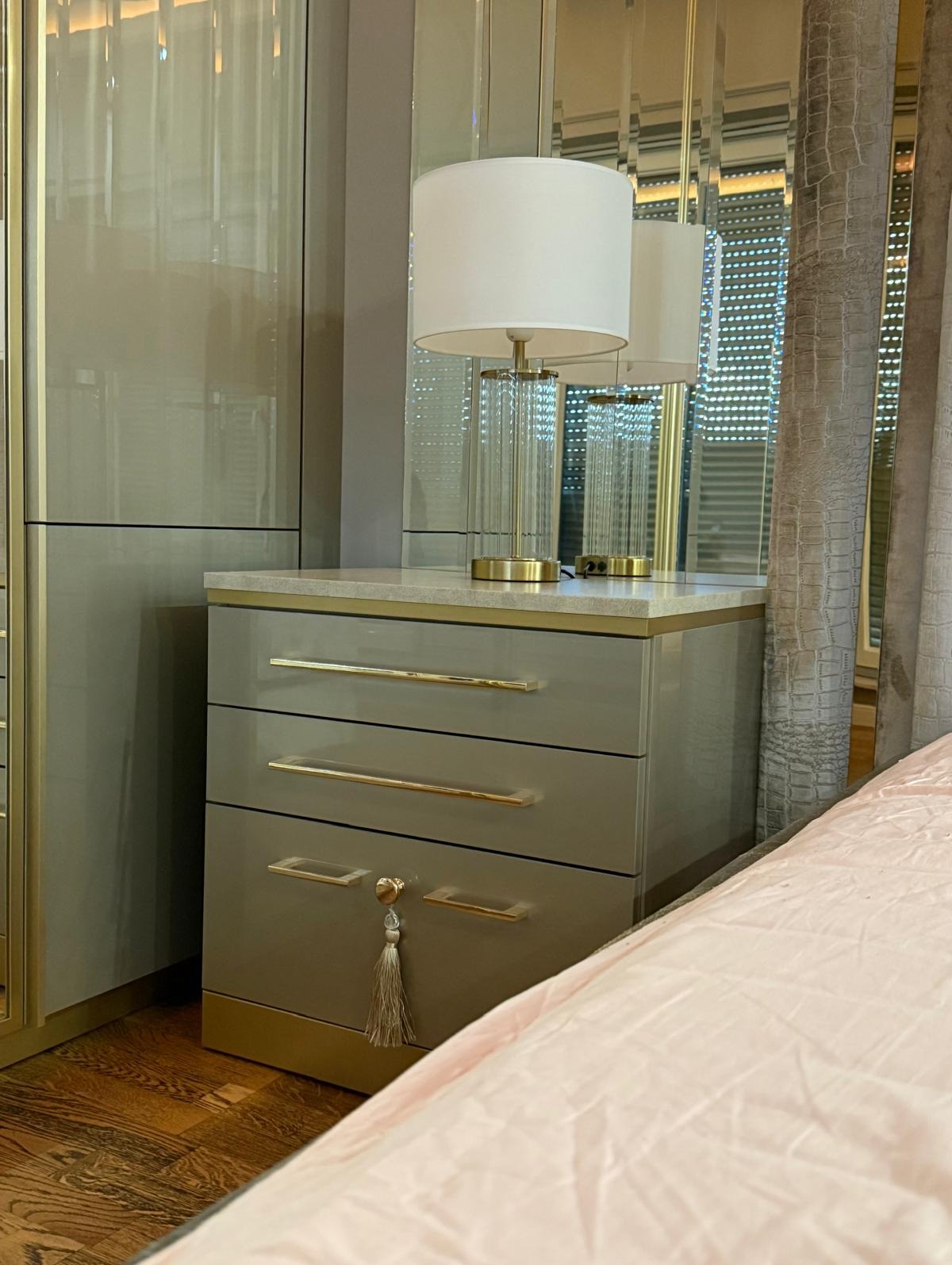 Szara szafka nocna z trzema szufladami i złotymi uchwytami, zwieńczona marmurowym blatem, na którym stoi szklana lampa z białym abażurem. Na jednej z szuflad wisi ozdobny chwost.