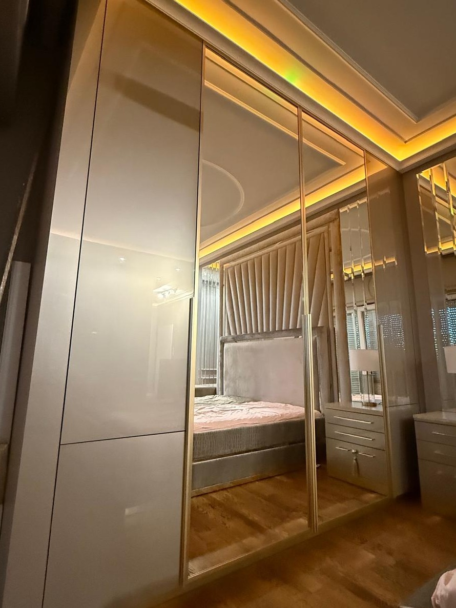 Nowoczesna szafa z przesuwnymi lustrzanymi drzwiami w sypialni, z widocznym łóżkiem z tapicerowanym zagłówkiem i oświetleniem LED na suficie.