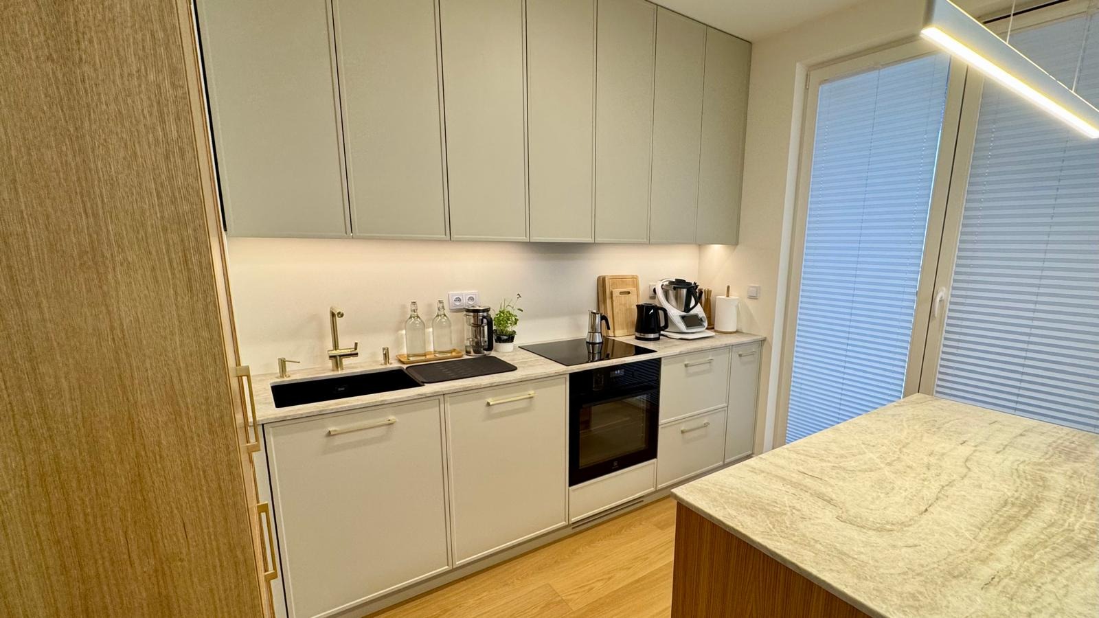 Nowoczesna kuchnia w odcieniach beżu i szarości z drewnianymi akcentami, czarnym zlewem i blatem z marmurkowym wzorem, oświetlona ledowym panelem.