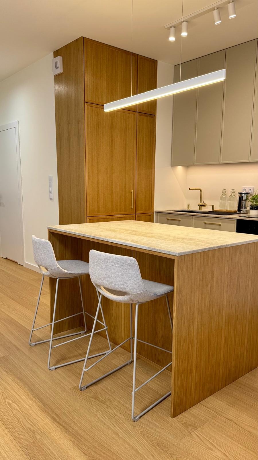 Nowoczesna kuchnia z drewnianą zabudową, wyspą z blatem w odcieniach beżu i dwoma szarymi krzesłami barowymi na metalowych nogach, oświetlona podłużną lampą LED.