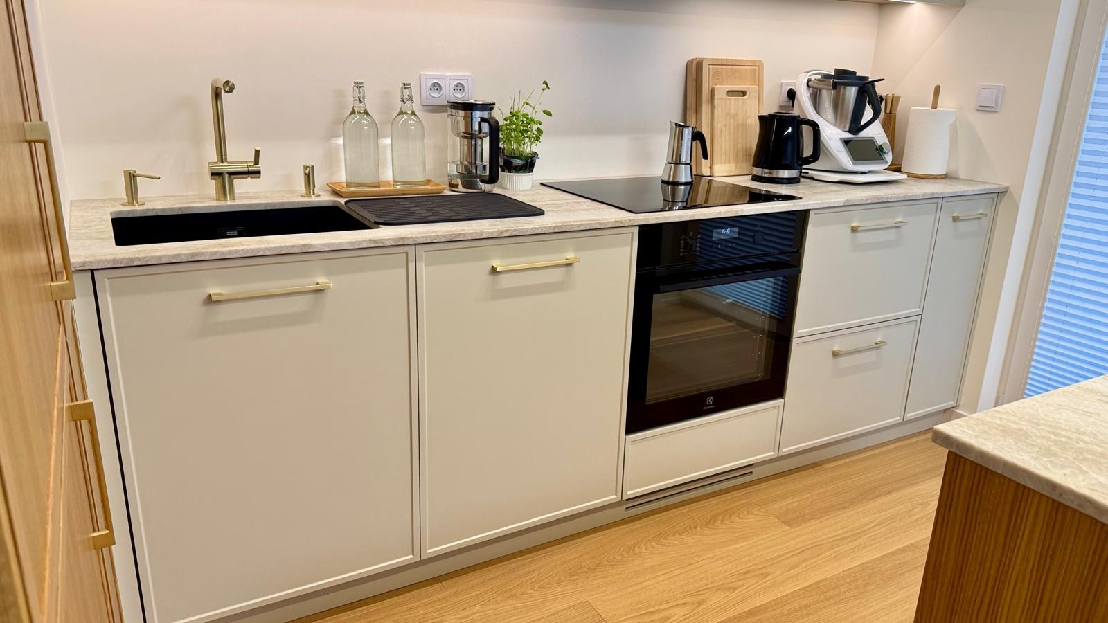 Minimalistyczna kuchnia z beżowymi frontami, złotymi uchwytami i czarnym zlewem. Na blacie ekspres do kawy, płyta indukcyjna i piekarnik Electrolux. Podłoga drewniana.