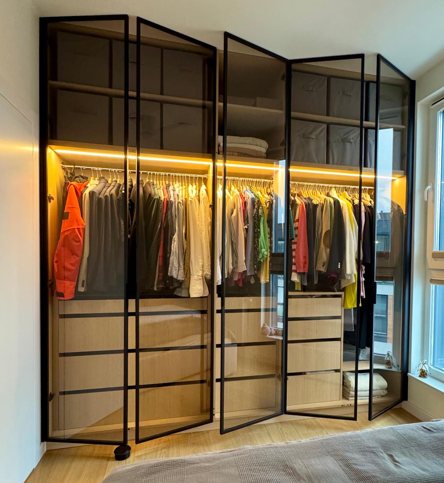 Nowoczesna szafa wnękowa z przesuwnymi drzwiami ze szkła w czarnej ramie, oświetlona listwą LED, z widoczną zawartością: ubrania na wieszakach i pudełka na półkach.