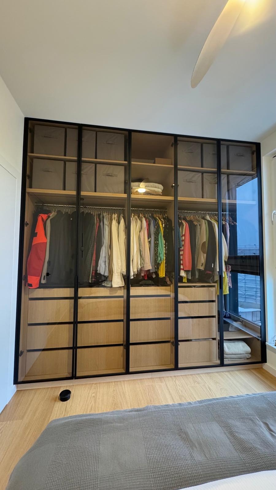 Duża szafa z przesuwnymi, przeszklonymi drzwiami w czarnych ramach, z widoczną zawartością: ubrania na wieszakach, pudełka i ręczniki na półkach, oraz szuflady z jasnego drewna.