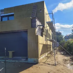 Dom w trakcie ocieplania: elewacja pokryta żółtą wełną mineralną, widoczne rusztowania i drewniana drabina oparta o ścianę, okno z szarą ramą, fragment garażu z czarną bramą, ziemia wokół budynku.