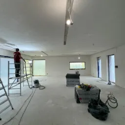 Pracownik na rusztowaniu szlifuje świeżo położone tynki maszynowe w przestronnym pomieszczeniu, widoczne okna zabezpieczone folią, materiały budowlane i odkurzacz przemysłowy.