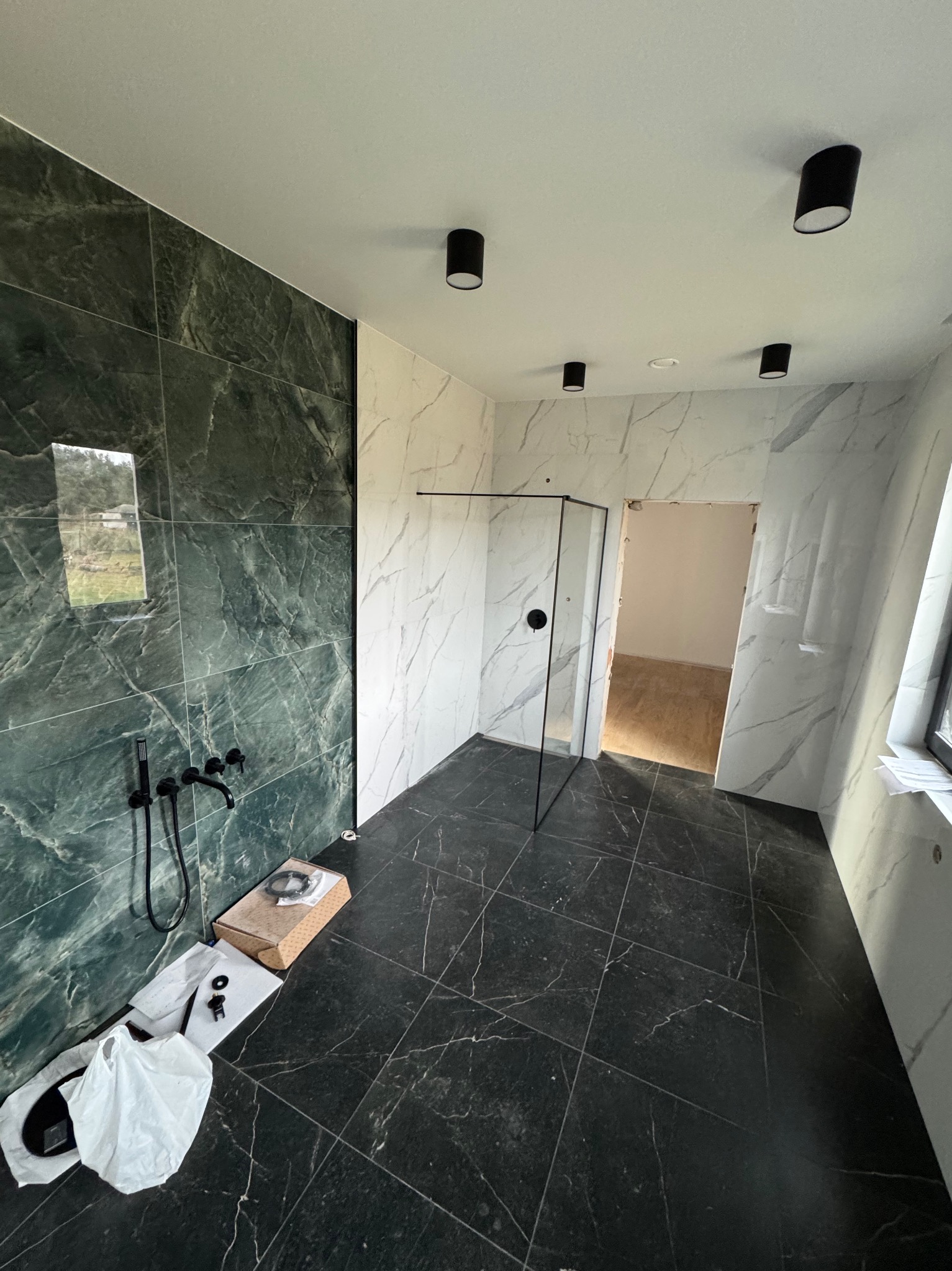 Nowoczesna łazienka z czarną armaturą i kabiną prysznicową typu walk-in. Ściany wyłożone płytkami imitującymi marmur w odcieniach zieleni i bieli, czarna podłoga.