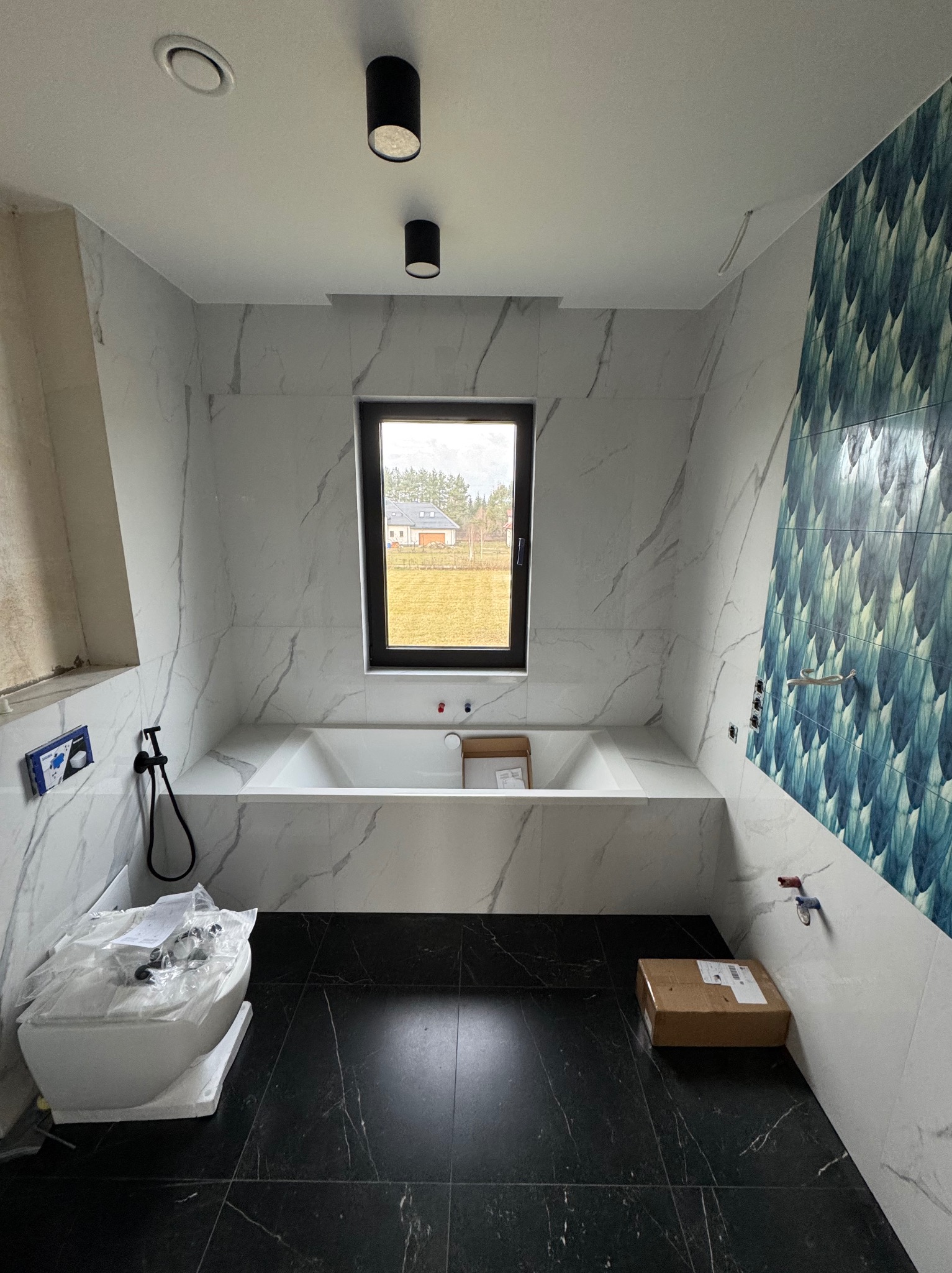Nowoczesna łazienka w trakcie wykańczania: białe marmurowe płytki, czarna podłoga, wanna pod oknem z widokiem na dom. Elementy instalacji widoczne.