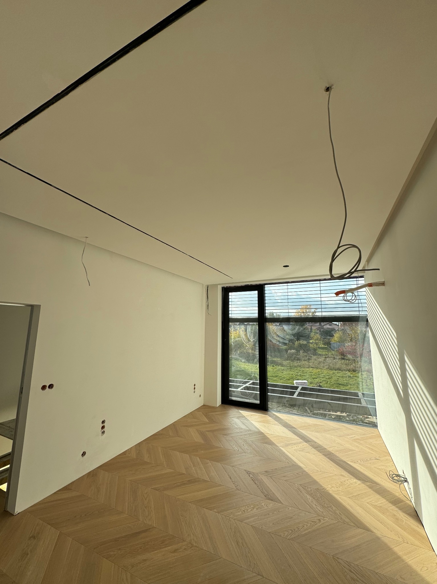 Wnętrze w stanie deweloperskim: parkiet w jodełkę, białe ściany z puszkami elektrycznymi, duże okno z widokiem na zieleń. Wystające kable elektryczne z sufitu.