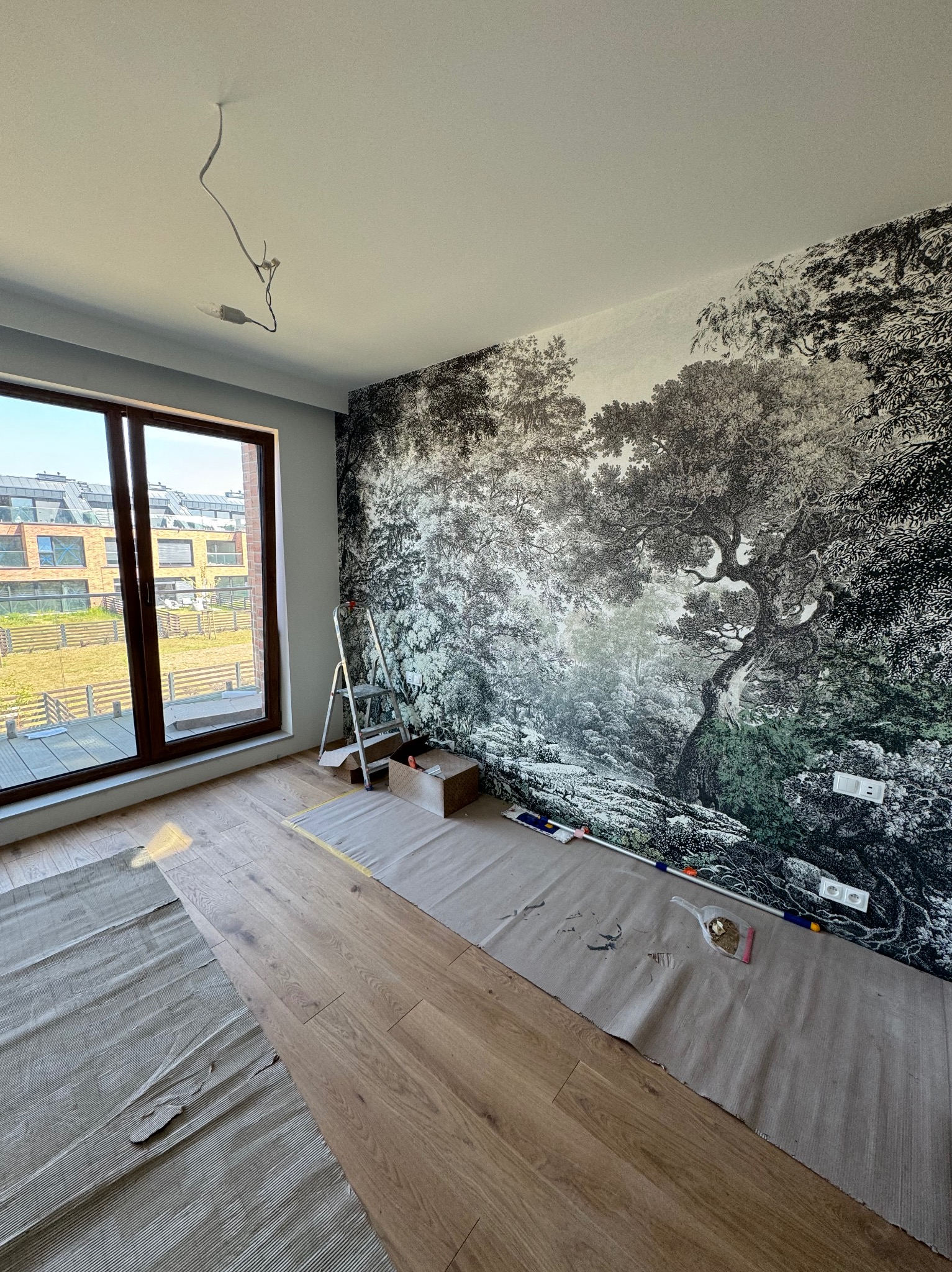 Pokój z fototapetą leśną w trakcie montażu: drabina, akcesoria malarskie i zabezpieczona podłoga z desek. Widok na osiedle przez okno balkonowe.