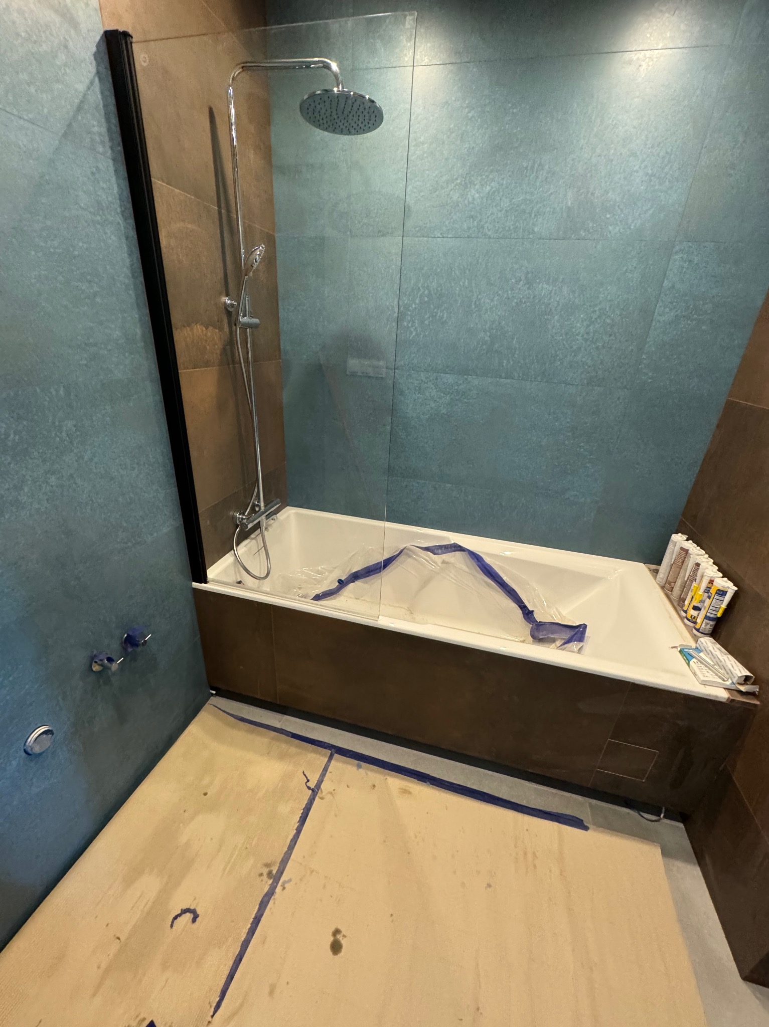 Remont łazienki w toku: zabezpieczona wanna folią, widoczne płytki w odcieniach brązu i turkusu, przygotowane silikony. Podłoga zabezpieczona kartonem.