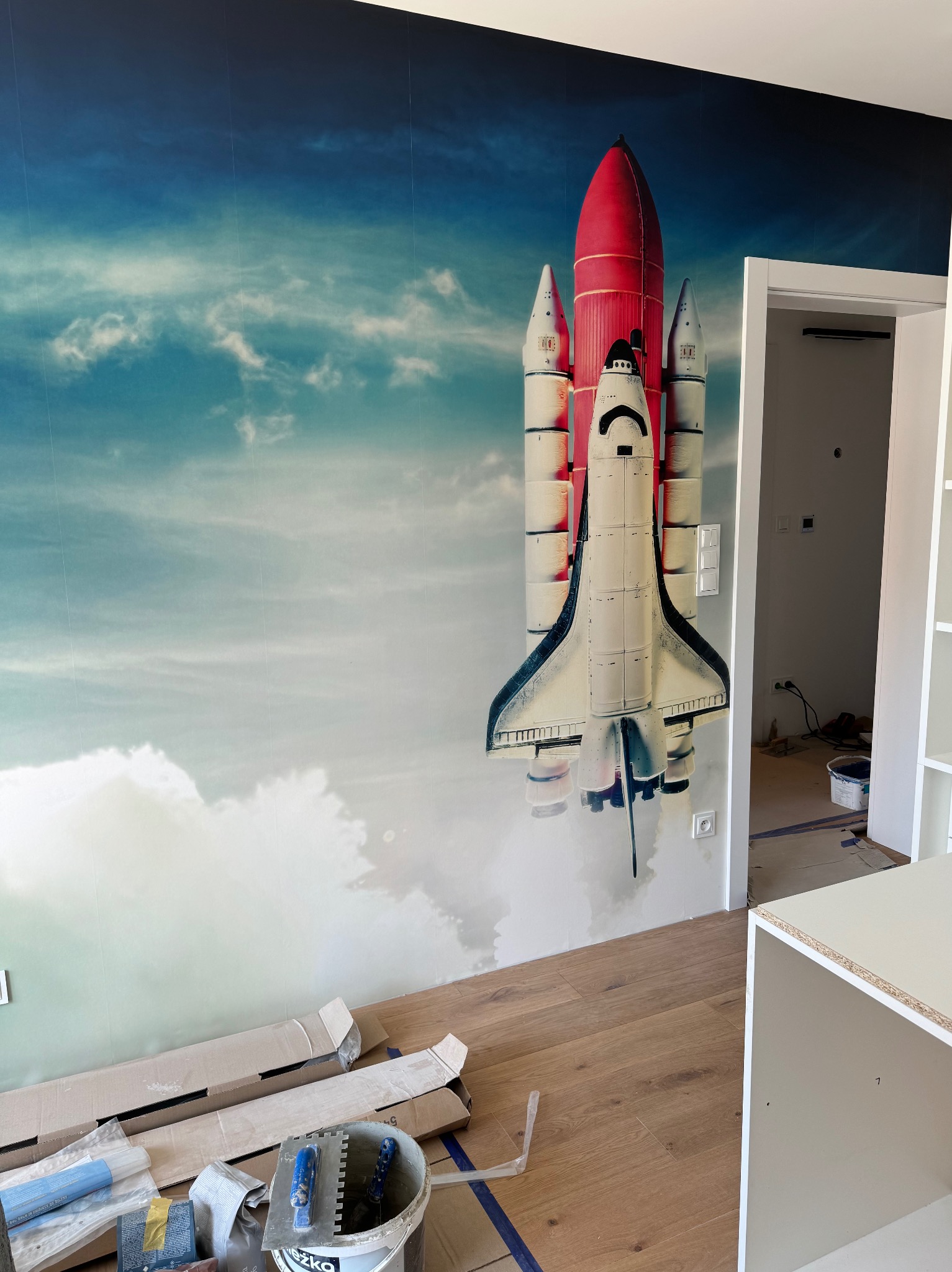 Pokój po remoncie, ściana z fototapetą z motywem rakiety kosmicznej na tle błękitnego nieba. Na podłodze widoczne narzędzia i materiały malarskie.