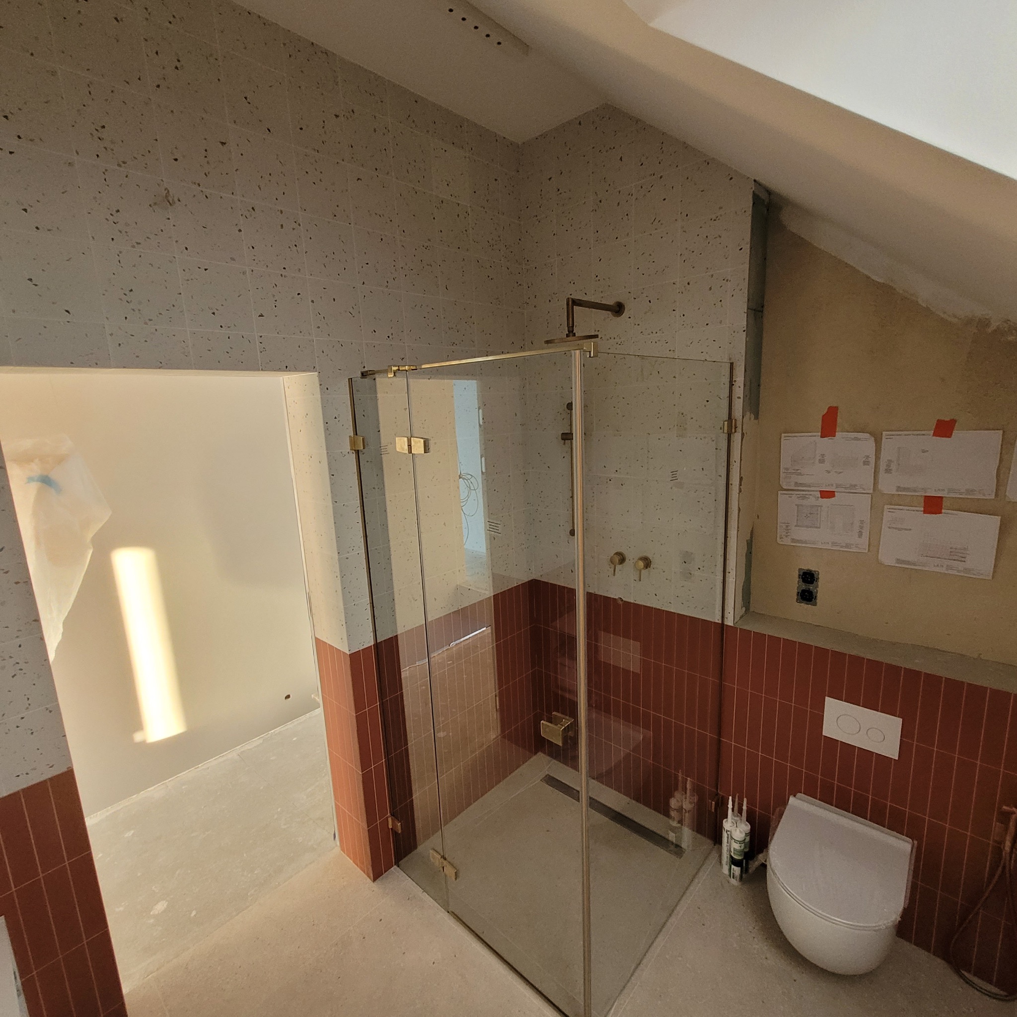 Nowoczesna kabina prysznicowa ze złotymi okuciami w łazience z beżowo-ceglastą mozaiką, widok na częściowo wykończone wnętrze i poddasze ze skośnym sufitem.