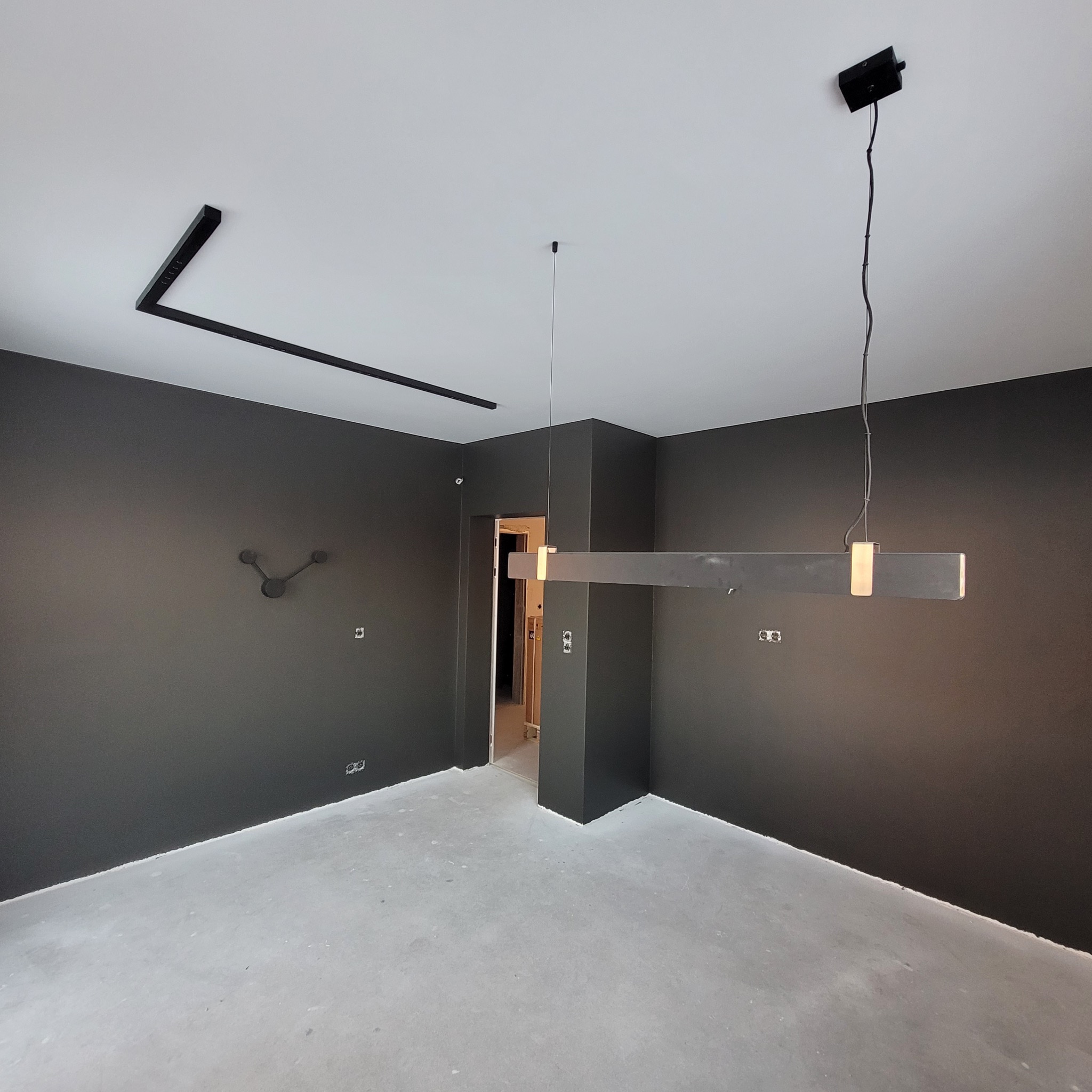 Surowe wnętrze z szarymi ścianami i betonową podłogą. Minimalistyczne oświetlenie sufitowe, widoczne gniazdka elektryczne i zegar na ścianie. Drzwi do innego pomieszczenia.