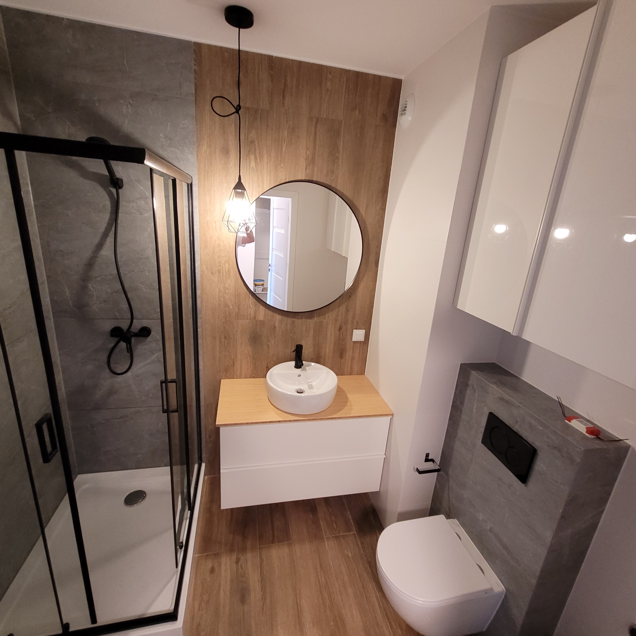 Nowoczesna łazienka z kabiną prysznicową typu walk-in, okrągłym lustrem i drewnopodobnymi płytkami. Umywalka nablatowa i wisząca toaleta.