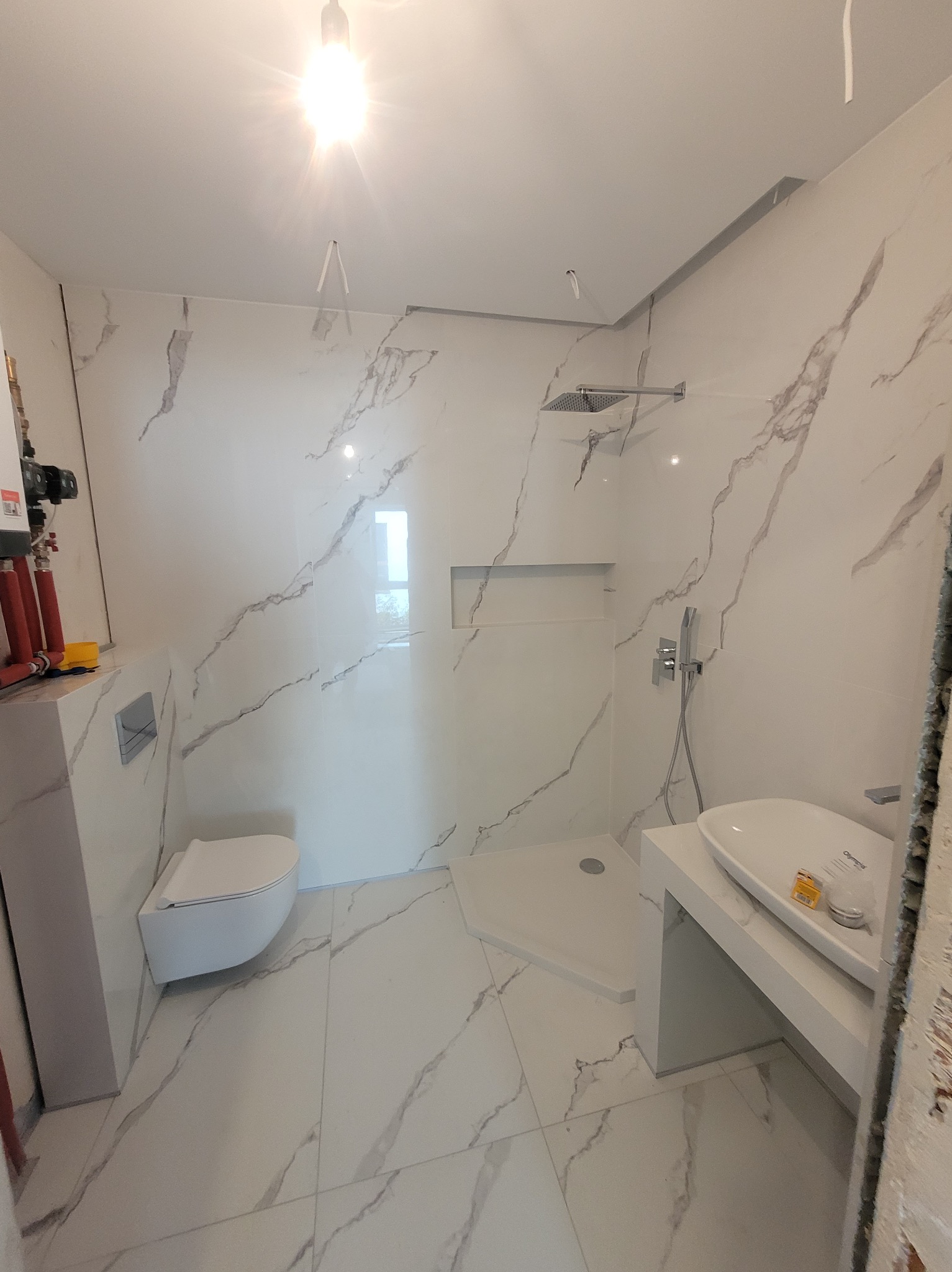 Wykończona łazienka z białymi płytkami imitującymi marmur, wisząca toaleta, kabina prysznicowa typu walk-in i umywalka na blacie. Widoczne przyłącza wodne.