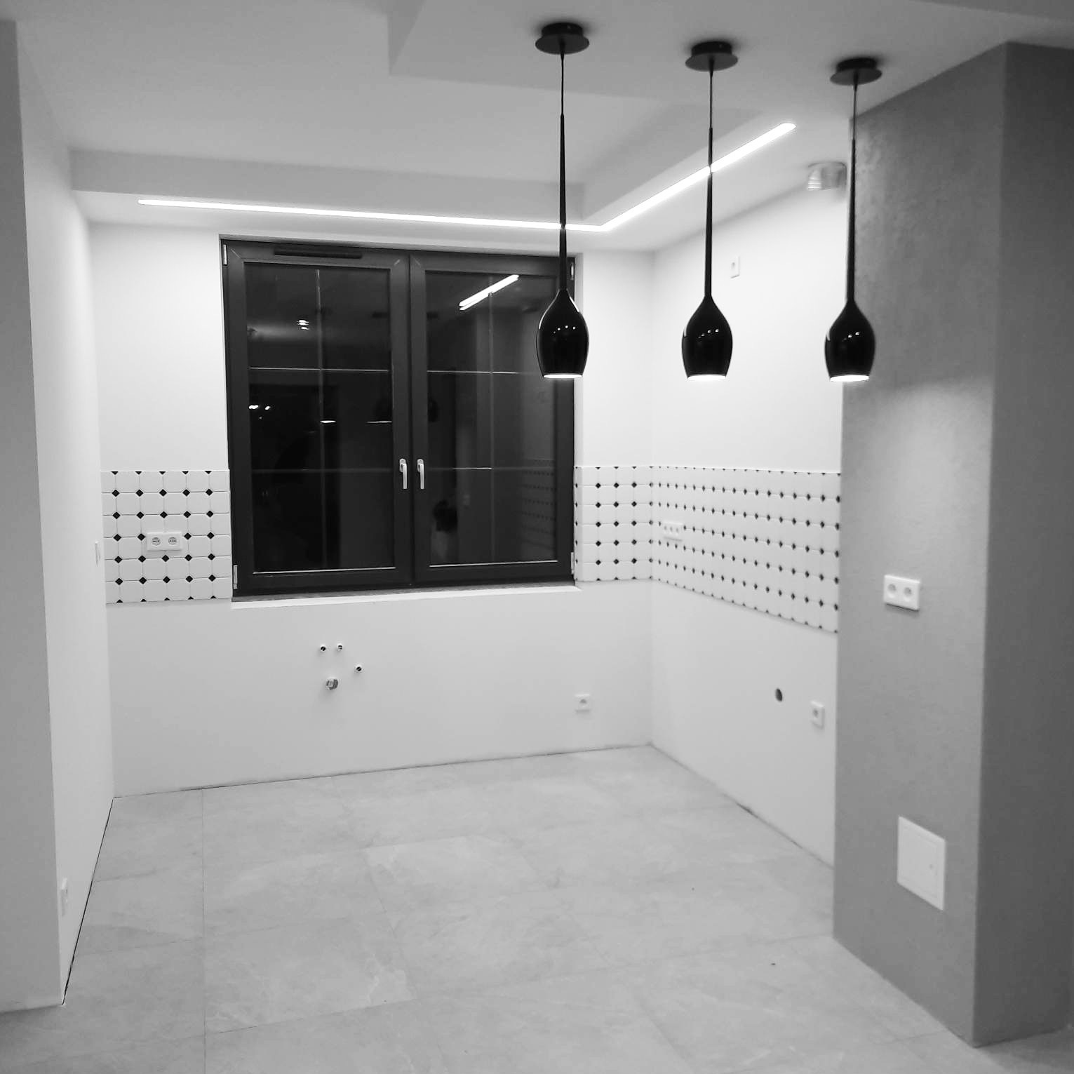 Czarno-białe zdjęcie wnętrza kuchni w trakcie remontu: okno, płytki na ścianie, trzy lampy wiszące. Widoczne instalacje wodne i elektryczne.