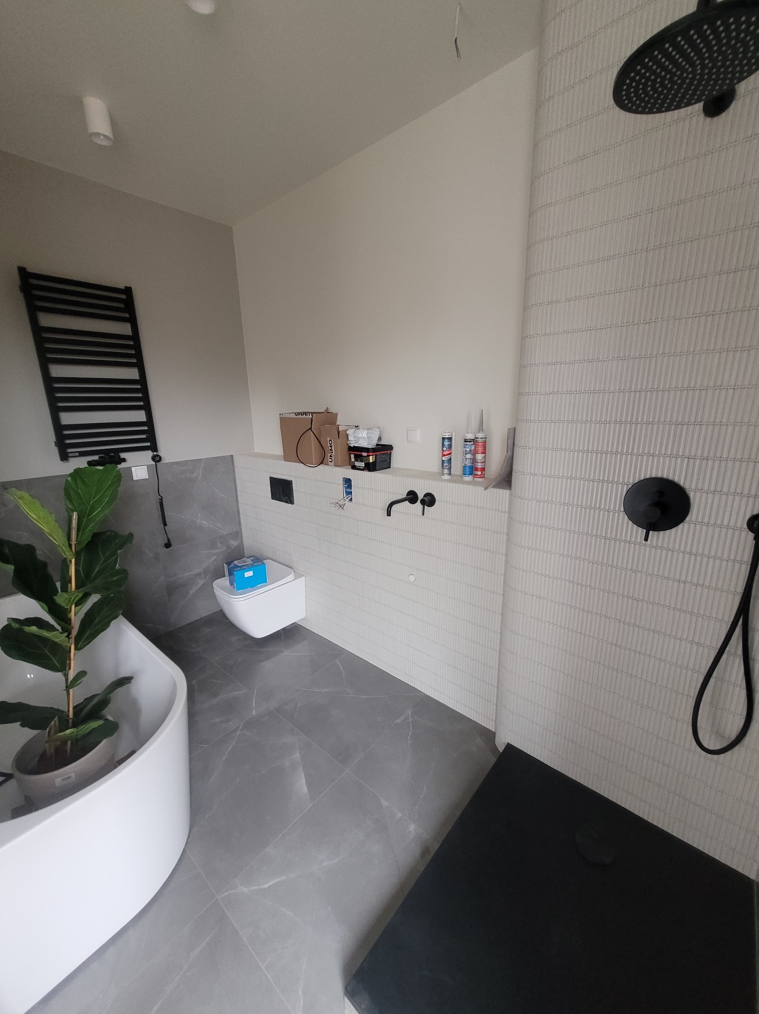 Nowoczesna łazienka w trakcie wykańczania: biała wanna, szara płytka podłogowa imitująca marmur, toaleta podwieszana i czarna armatura prysznicowa.