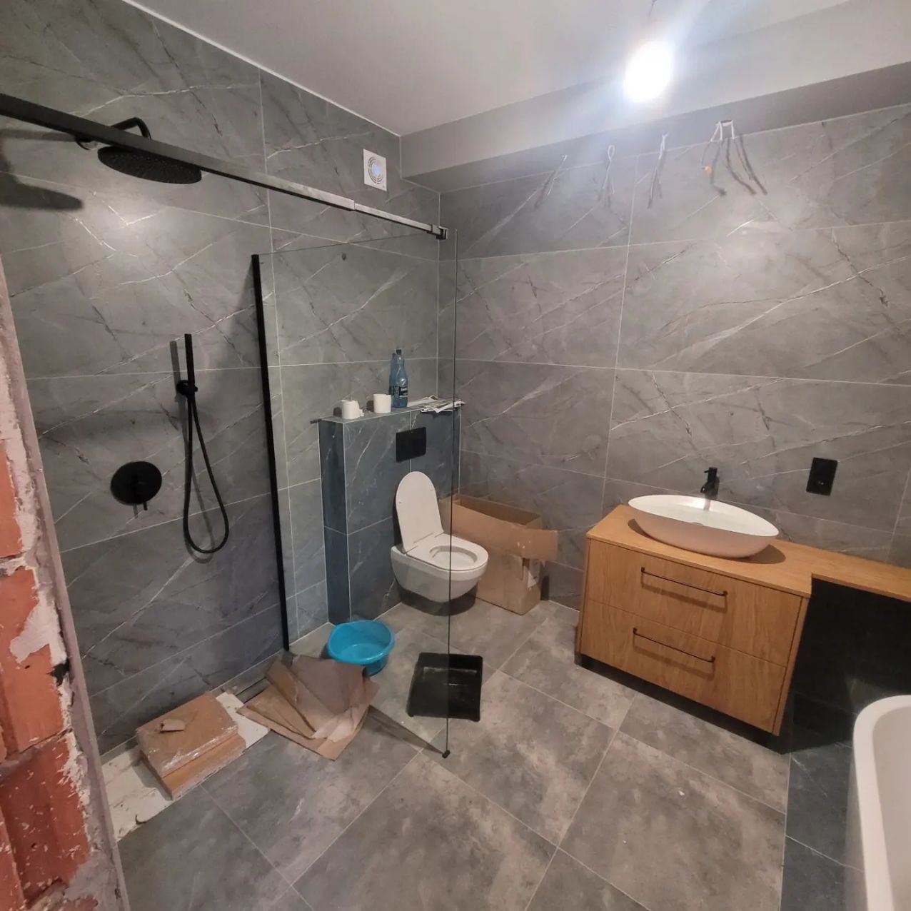Wykończona łazienka z szarymi płytkami imitującymi marmur, kabiną prysznicową typu walk-in z czarną armaturą, umywalką nablatową i drewnianą szafką. Widoczne elementy po remoncie.