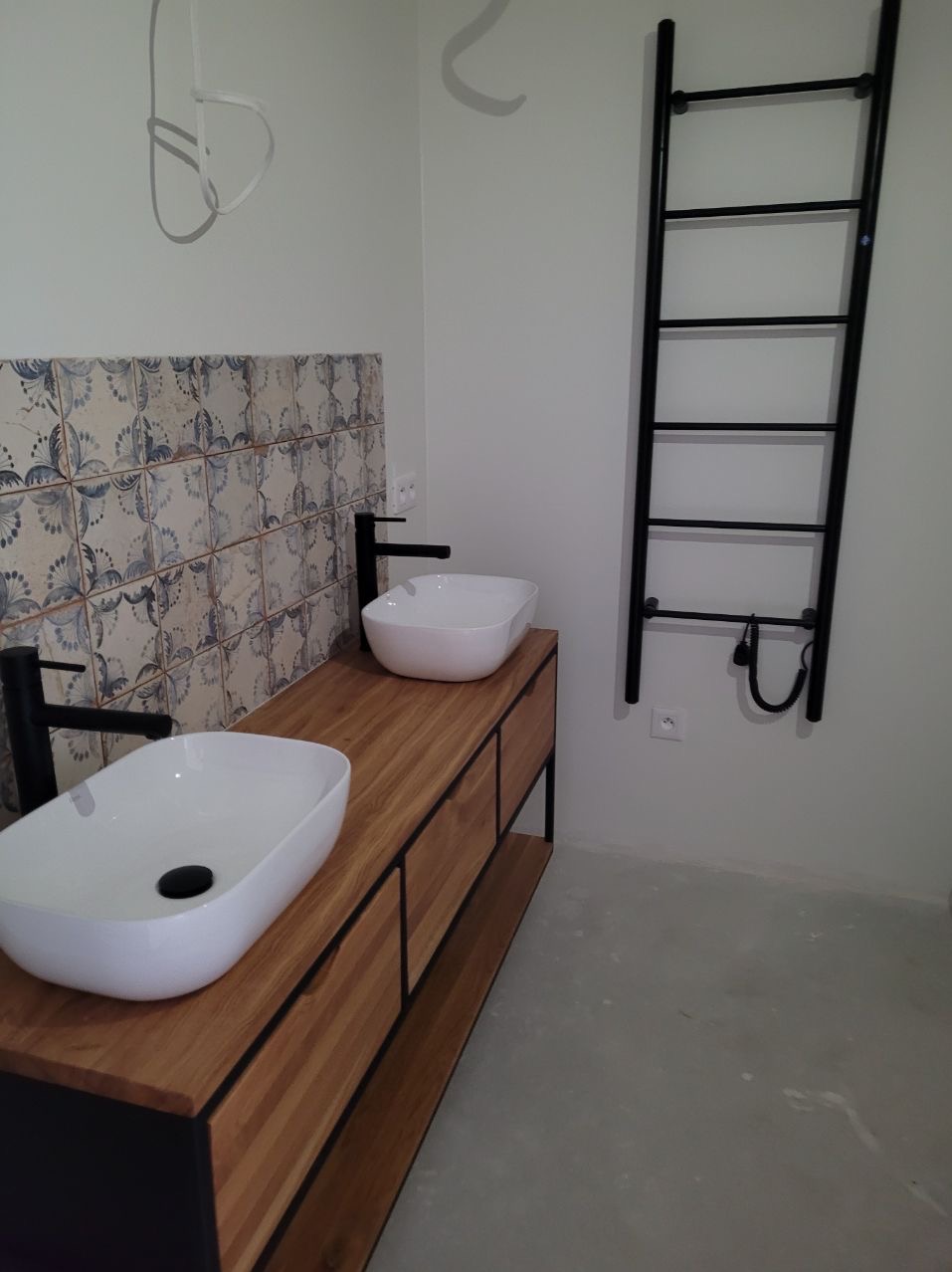 Nowoczesna łazienka z drewnianą szafką pod umywalki, wzorzystą glazurą i czarnym grzejnikiem drabinkowym. Minimalistyczny design, białe umywalki nablatowe.