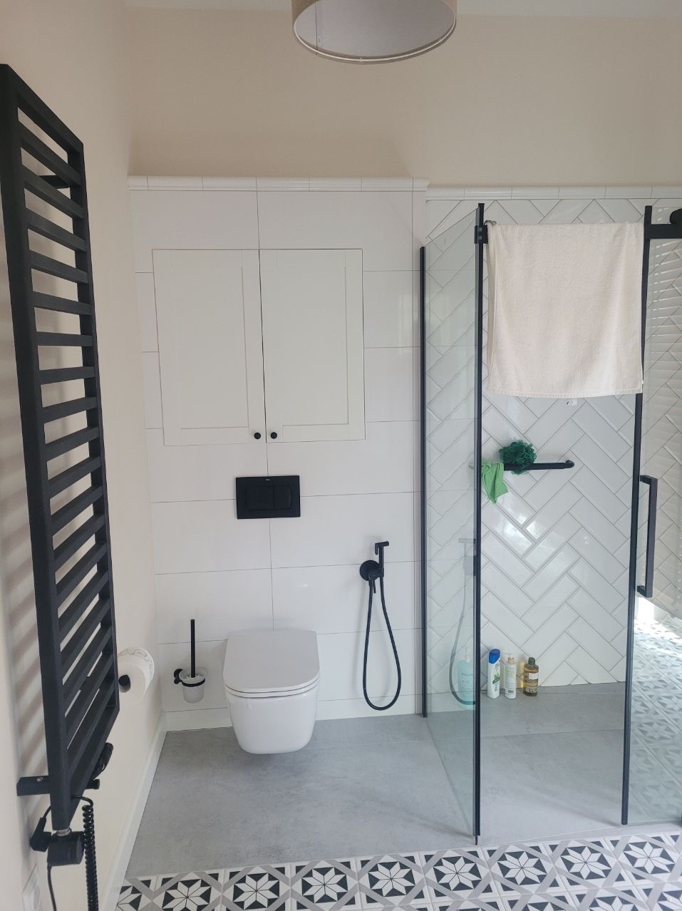 Nowoczesna łazienka po remoncie: białe płytki, kabina prysznicowa ze szklaną ścianą, czarna armatura i grzejnik drabinkowy. Wzór na podłodze.