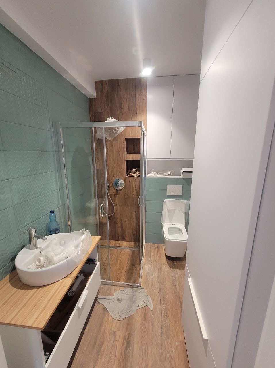 Nowoczesna łazienka w trakcie wykańczania: kabina prysznicowa z drewnianą ścianą, umywalka na szafce z jasnego drewna, toaleta i turkusowe płytki na ścianach.