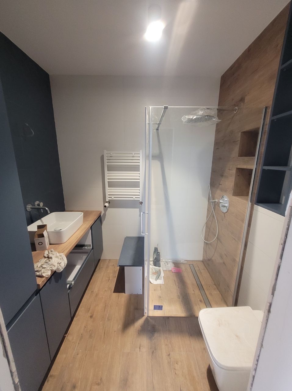 Nowoczesna łazienka po remoncie w Warszawie: kabina prysznicowa typu walk-in z odpływem liniowym, drewnopodobne płytki, szara zabudowa meblowa z umywalką.