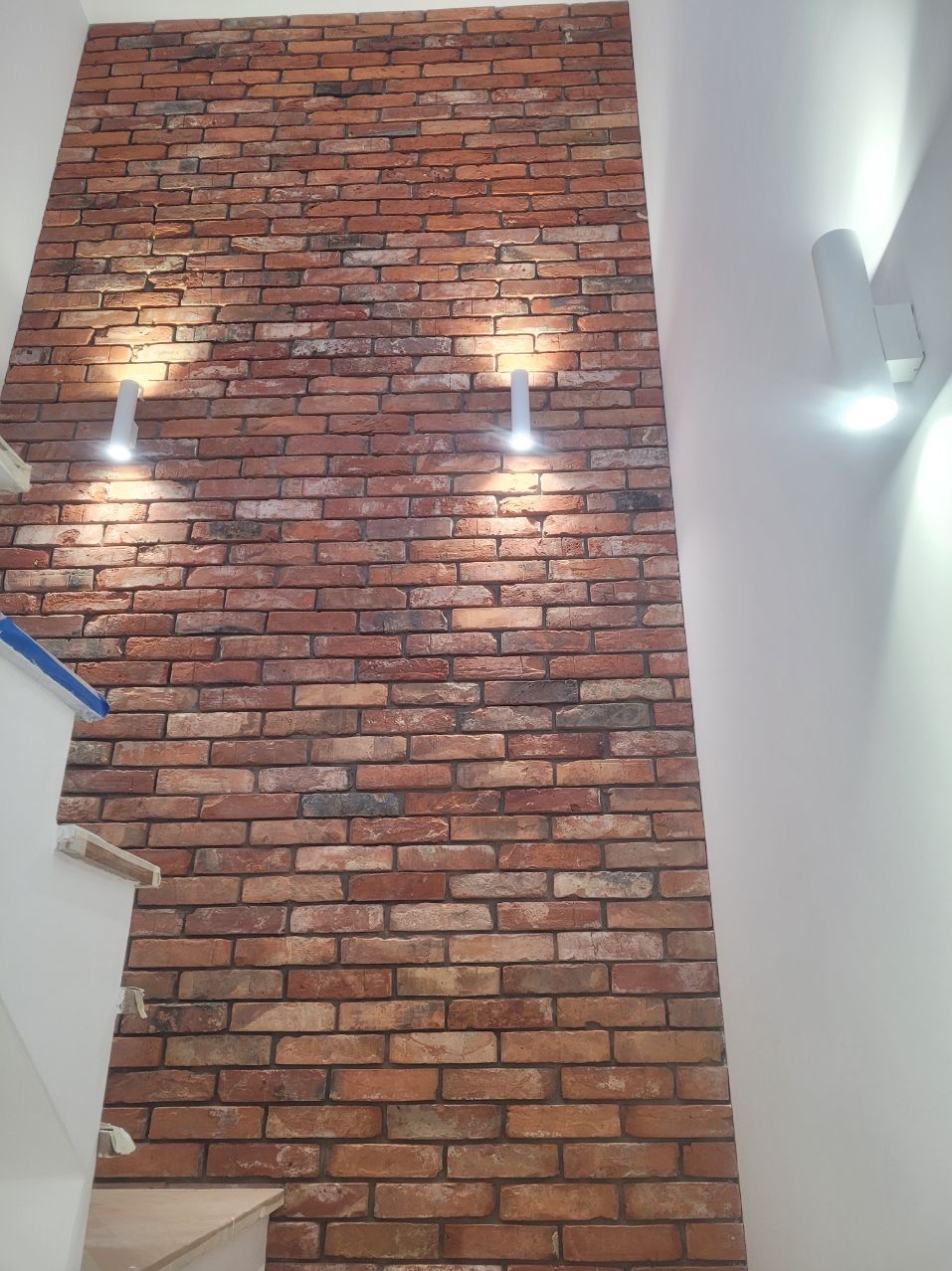 Ściana z cegły klinkierowej, oświetlona dwoma kinkietami, z fragmentem schodów po lewej stronie. Widoczne nieregularne spoiny i zróżnicowana kolorystyka cegieł.