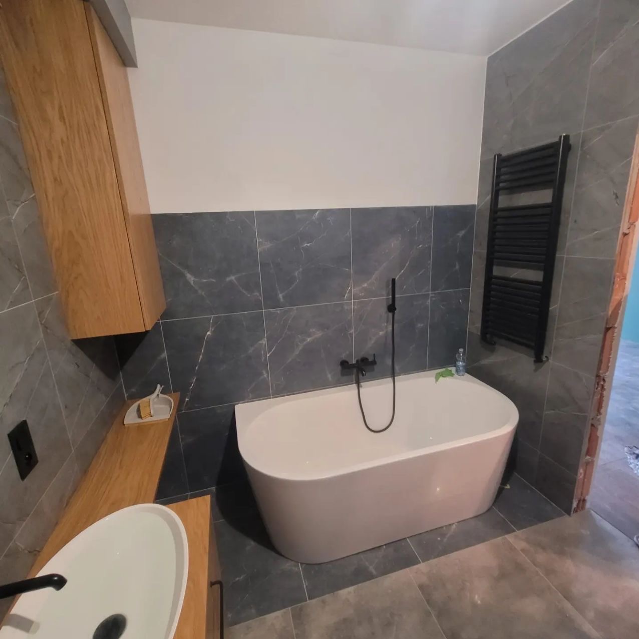 Nowoczesna łazienka z wanną wolnostojącą, szarą marmurową okładziną, drewnianą szafką pod umywalką i czarnym grzejnikiem drabinkowym. Minimalistyczny design.