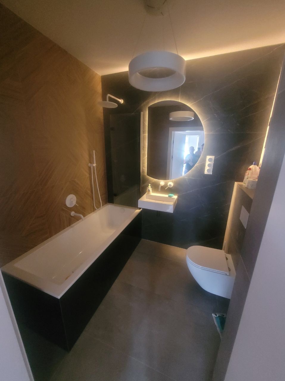 Nowoczesna łazienka z wanną w zabudowie, umywalką, podświetlanym lustrem i szarą podłogą. Wykończenie w odcieniach brązu i czerni. Widok na całe pomieszczenie.