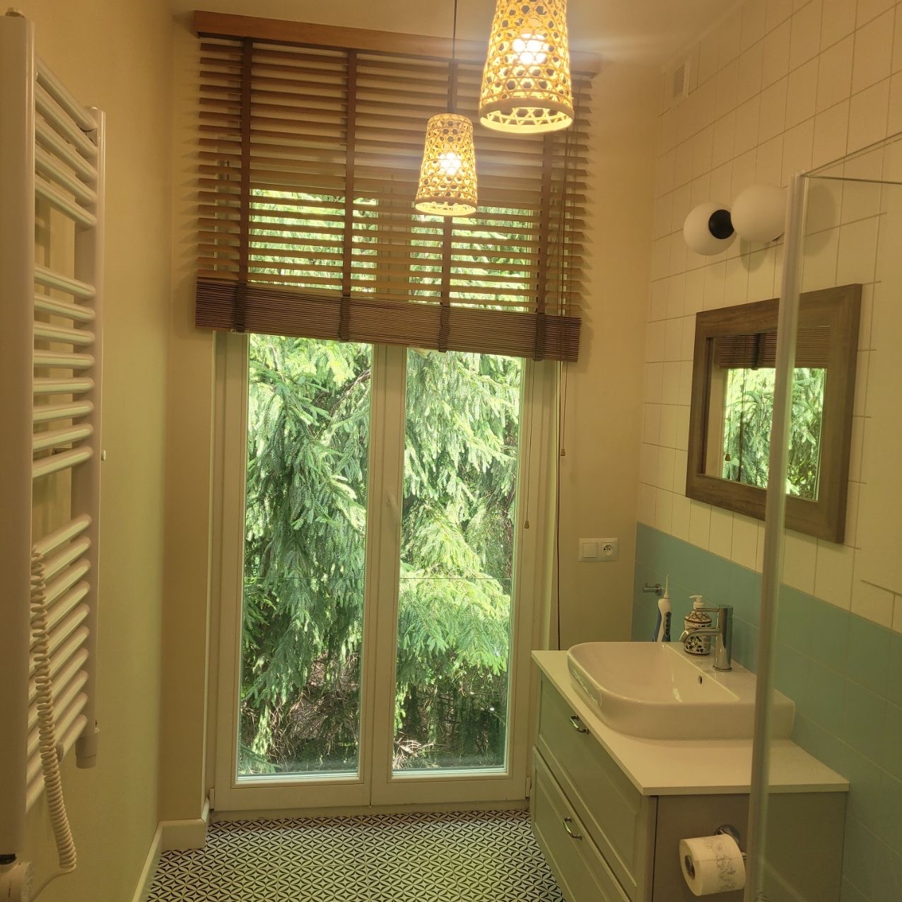 Jasna łazienka z oknem na zieleń, drewniane żaluzje i wzorzysta podłoga. Umywalka z szarą szafką, ozdobne lampy wiszące, lustro w drewnianej ramie.