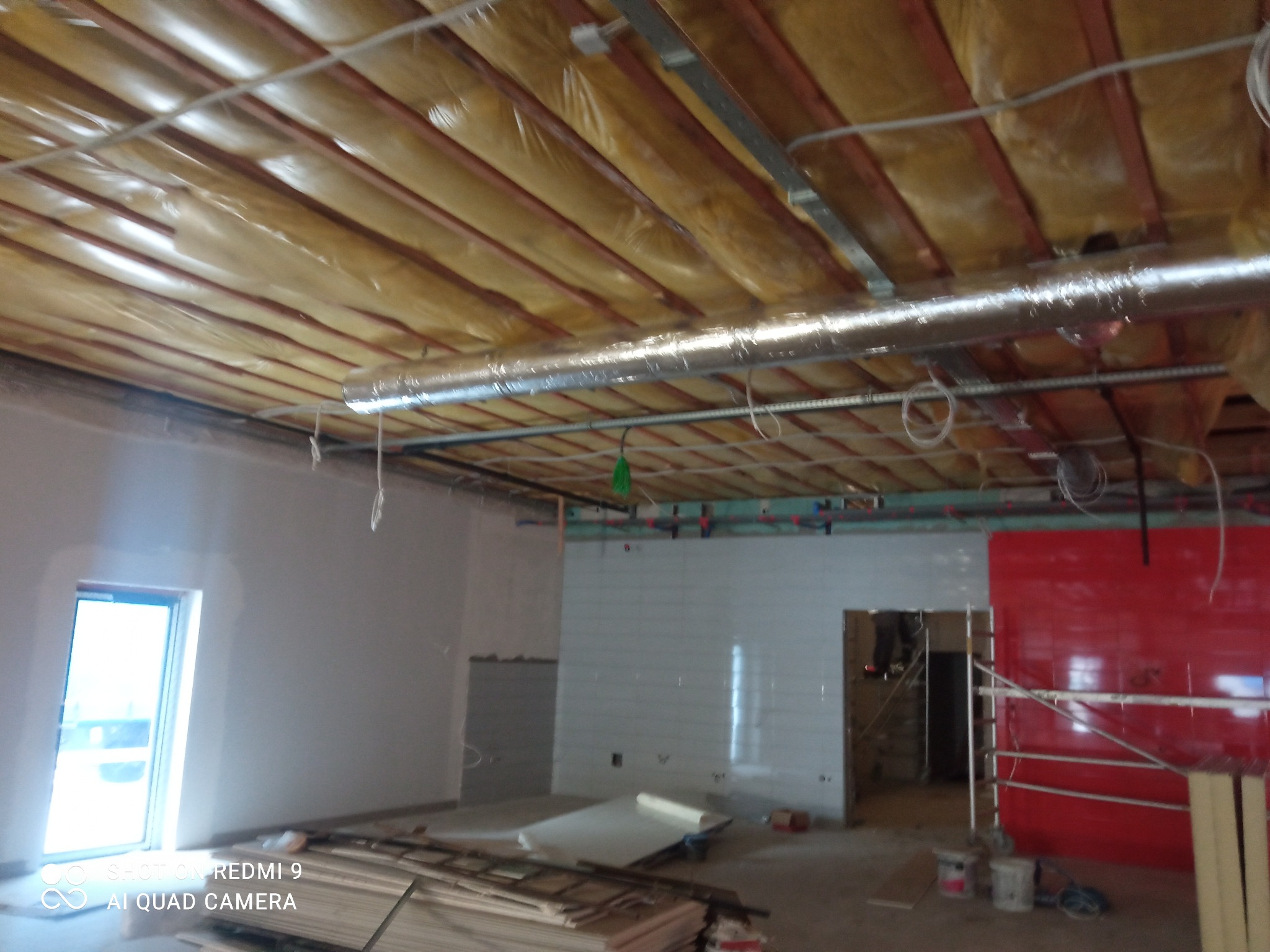 Wnętrze w trakcie prac wykończeniowych: ocieplony sufit, instalacje wentylacyjne, fragment ściany z białymi płytkami, czerwona ściana, okno z widokiem na zewnątrz, materiały budowlane na podłodze.