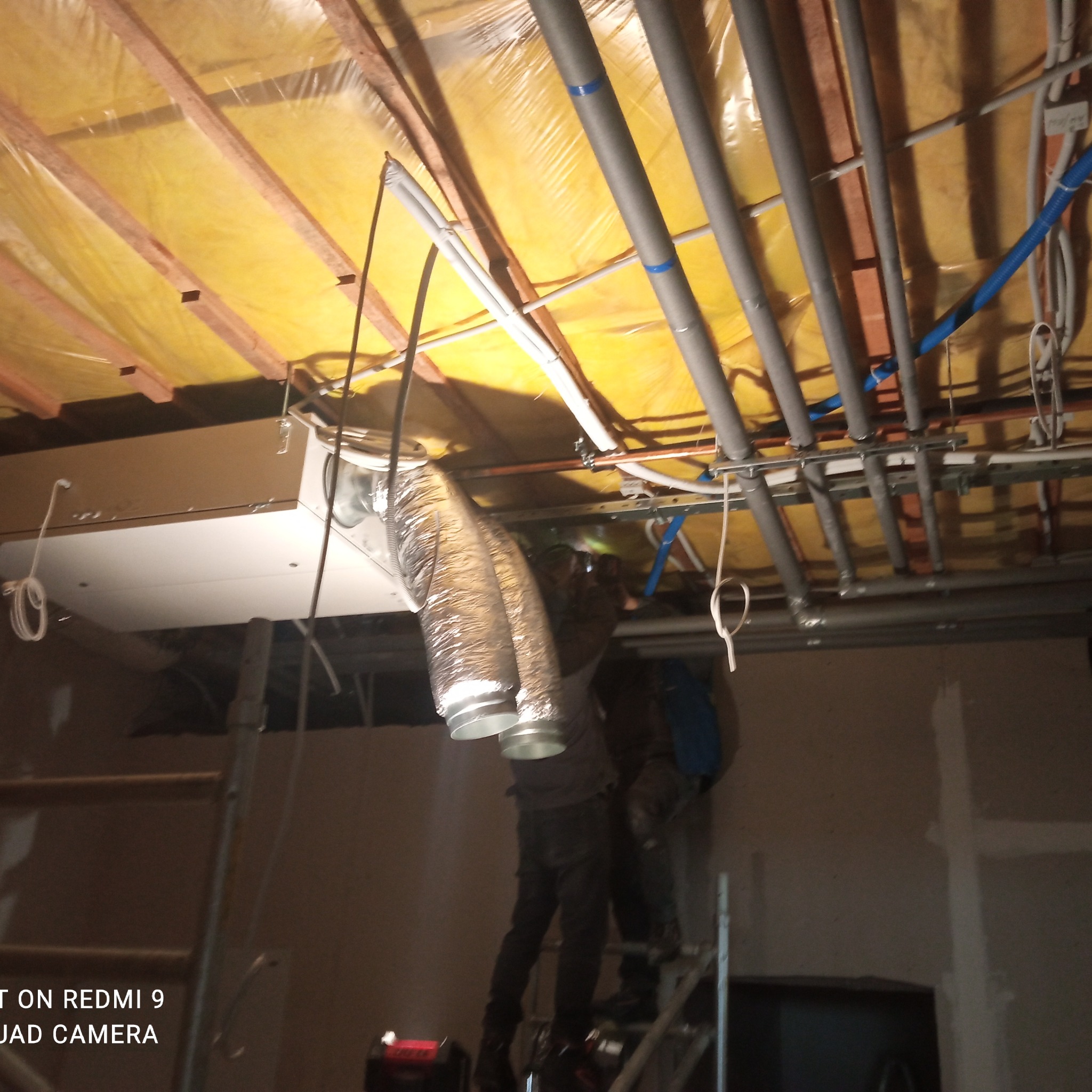 Montaż przewodów wentylacyjnych z folii aluminiowej pod żółtą izolacją stropu, widoczne rury i instalacje, pracownicy na drabinie w trakcie pracy.