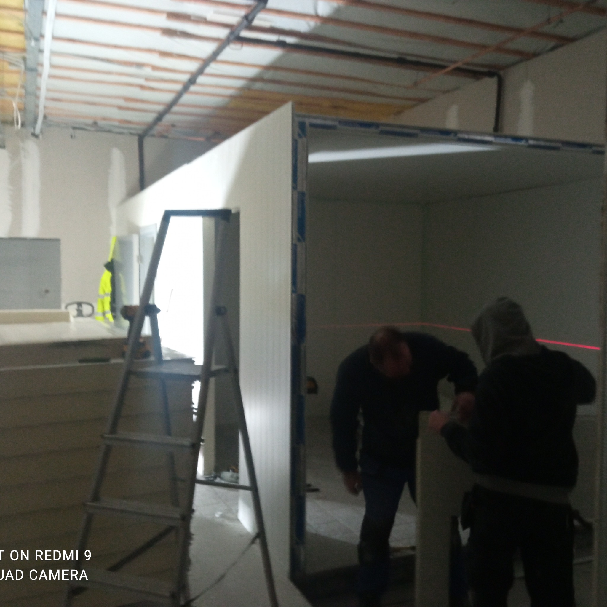 Montaż ścian działowych z użyciem lasera poziomującego w surowym stanie deweloperskim, widoczna drabina i pracownicy w odzieży roboczej.