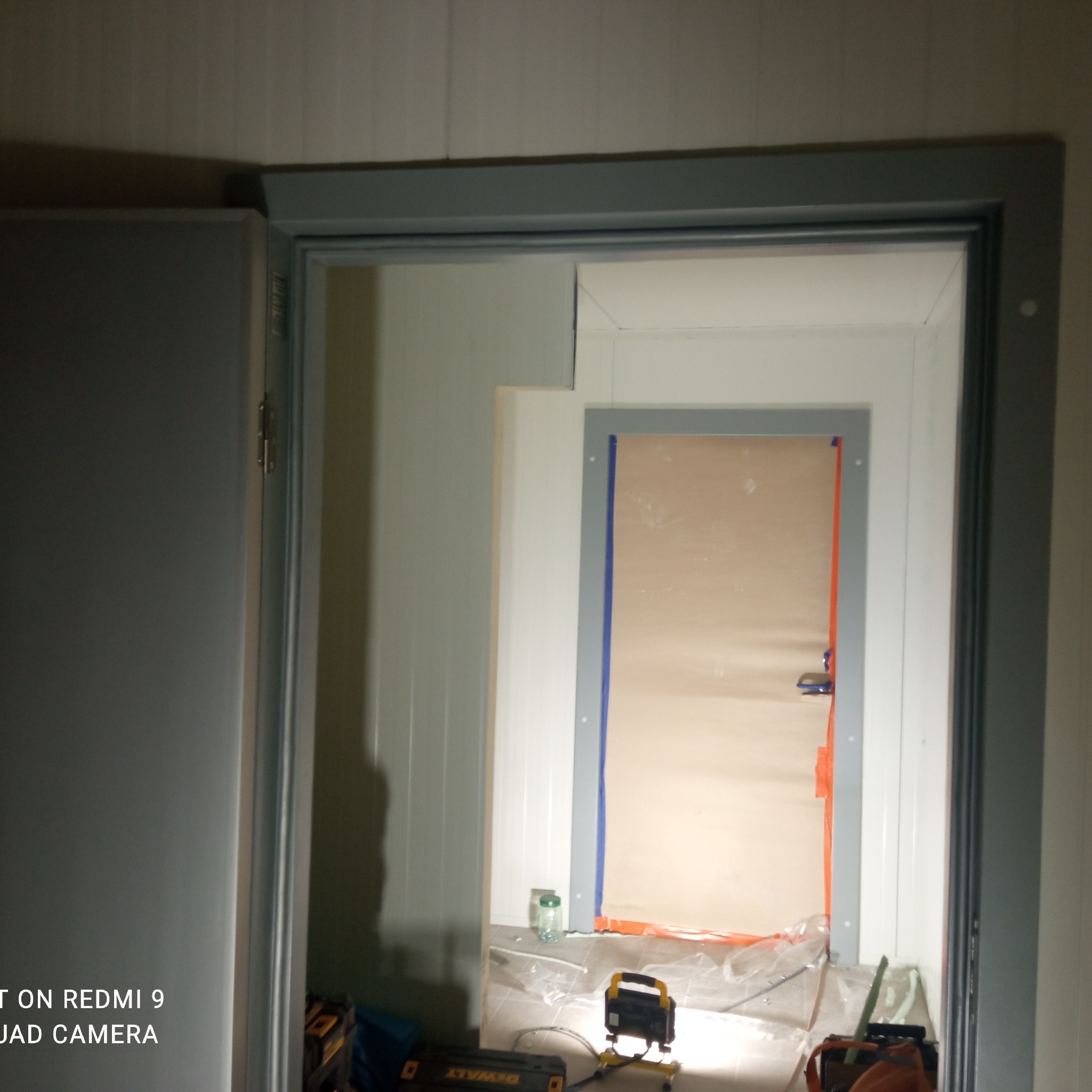 Montaż ościeżnicy metalowej w nowym budynku, widoczne narzędzia i lampa budowlana, zabezpieczenia folią i taśmą malarską.