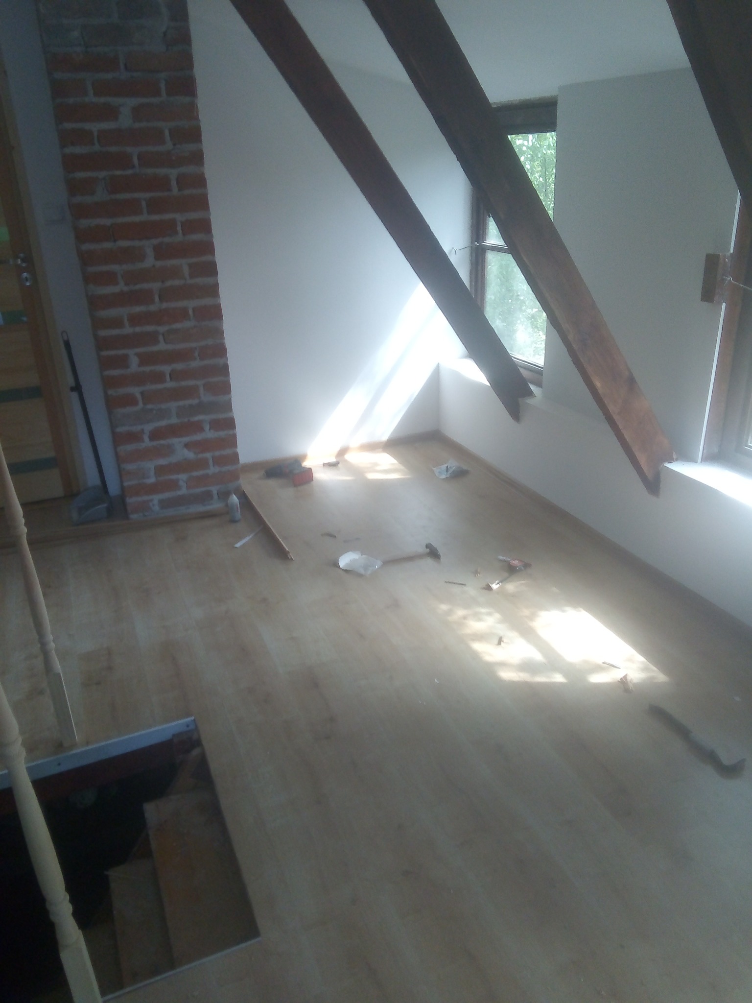 Poddasze w trakcie wykańczania: widoczne drewniane belki stropowe, fragment ściany z cegły, okno z widokiem na zieleń oraz rozłożone narzędzia na podłodze z paneli.